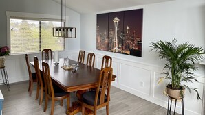 A dining room with a wooden table and chairs, a chandelier, a cityscape painting, and a potted plant. - Beautiful relaxing home 4BR/3BA near Light Rail (Seattle)