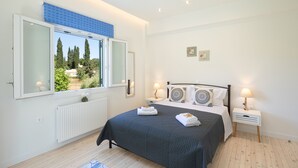 A bedroom with a large bed, a window with a view of trees, a bedside table with a lamp, and a radiator. - Villa Olga by Corfuescapes (Corfu)