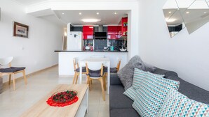 A modern living room with a sofa, a dining table with chairs, and a kitchen area with red cabinets and a white refrigerator.