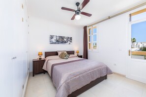 A bedroom with a bed, nightstands, a ceiling fan, a window with curtains, and a painting on the wall.