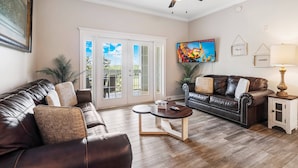 A living room with a brown leather sofa, a wooden coffee table, and a ceiling fan. - Heritage Crossing Magic | 3BR, 2 Bath Cozy Condo w/ Golf Course Views and Themed Room Near Disney (Kissimmee)