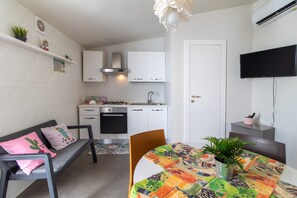 Cocina pequeña con gabinetes blancos, estufa y lavamanos. Hay una mesa de comedor con mantel colorido y una planta en maceta.