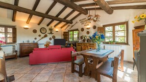 A rustic interior with exposed wooden beams, a red sofa, a wooden dining table, and a kitchen area with a window. - Villa "Stazzu La Piredda" with Garden & Wi-Fi (Arzachena)