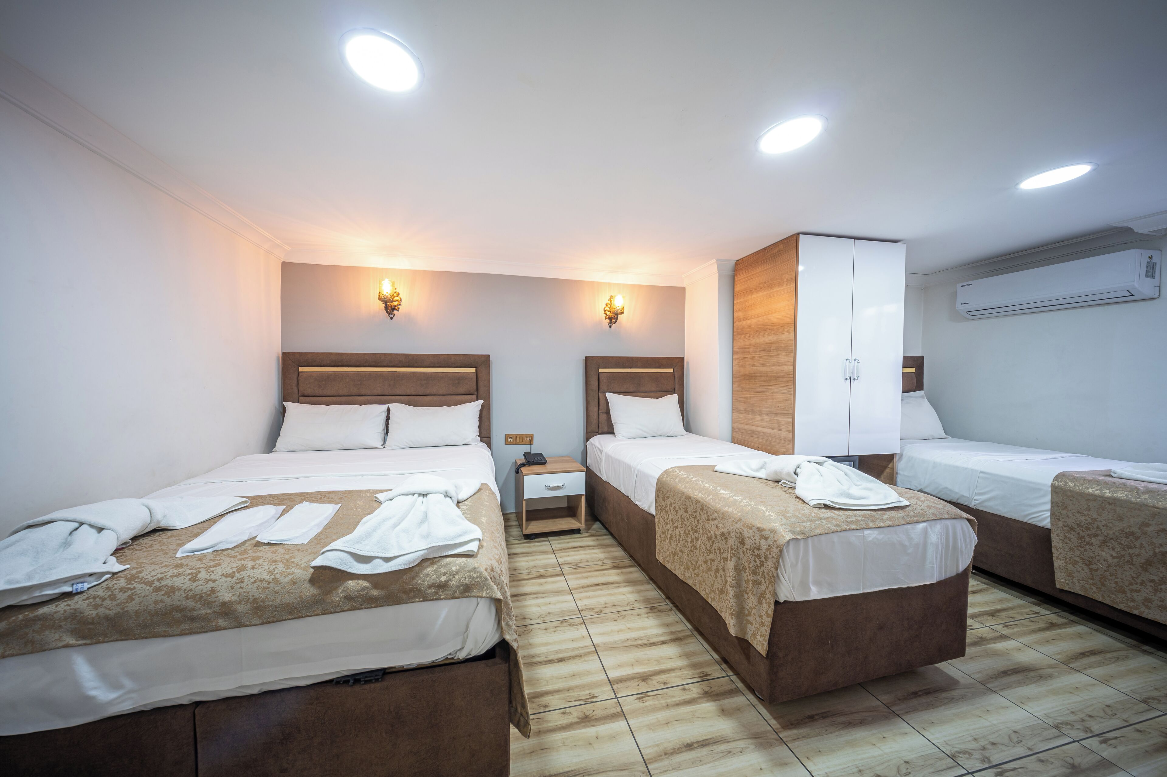 A hotel room with three beds, a wooden wardrobe, a nightstand, and an air conditioning unit.