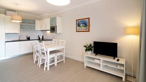 Una cuina moderna amb armaris blancs, una taula de menjador amb caires, un televisor de pantalla plana sobre un suport blanc i un quadre a la paret.