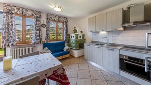 A modern kitchen with a built-in oven, a dining area with a table and a blue sofa, and a view of the outdoors through large windows.