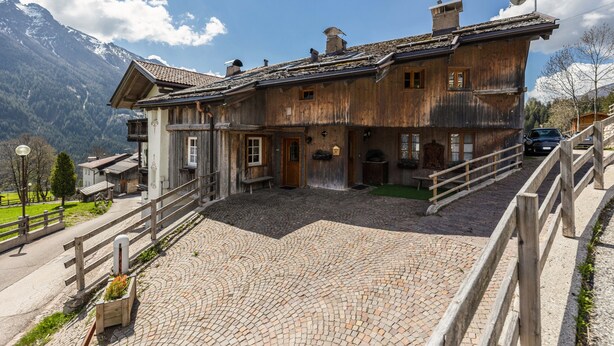 A wooden chalet with a stone courtyard, a wooden fence, and a mountain backdrop.