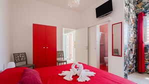 Une chambre d’hôtel avec une couvre-lit rouge, un téléviseur fixé au mur et une salle de bain visible à travers une porte ouverte.