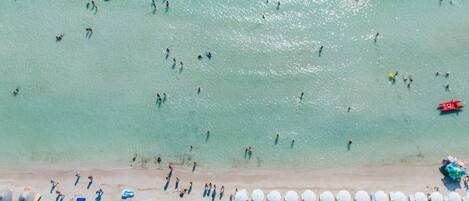 Vista aérea de una playa con gente nadando y tomando el sol, y filas de sombrillas y sillas de playa.