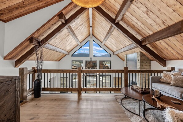 Un loft espacioso con techo alto, vigas de madera, un balcón con barandilla, un ventanal con vistas a la montaña y una zona de estar acogedora con sofá y mesa de centro.