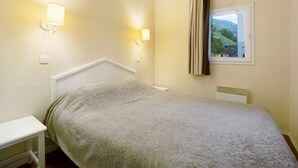 A bedroom with a bed, bedside table, wall-mounted light fixtures, a window with curtains, and a radiator. - Vacation Apartment "Ski-In/Ski-Out" Perfectly Located Year-Round (Valmeinier)