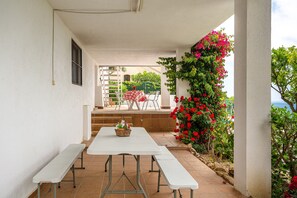 Een overdekt terras met witte tafels en banken, een mand met bloemen, en uitzicht op een tuin met rode bloemen.
