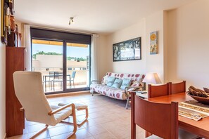 A living room with a sofa, a dining table, a chair, and a view through a sliding glass door.