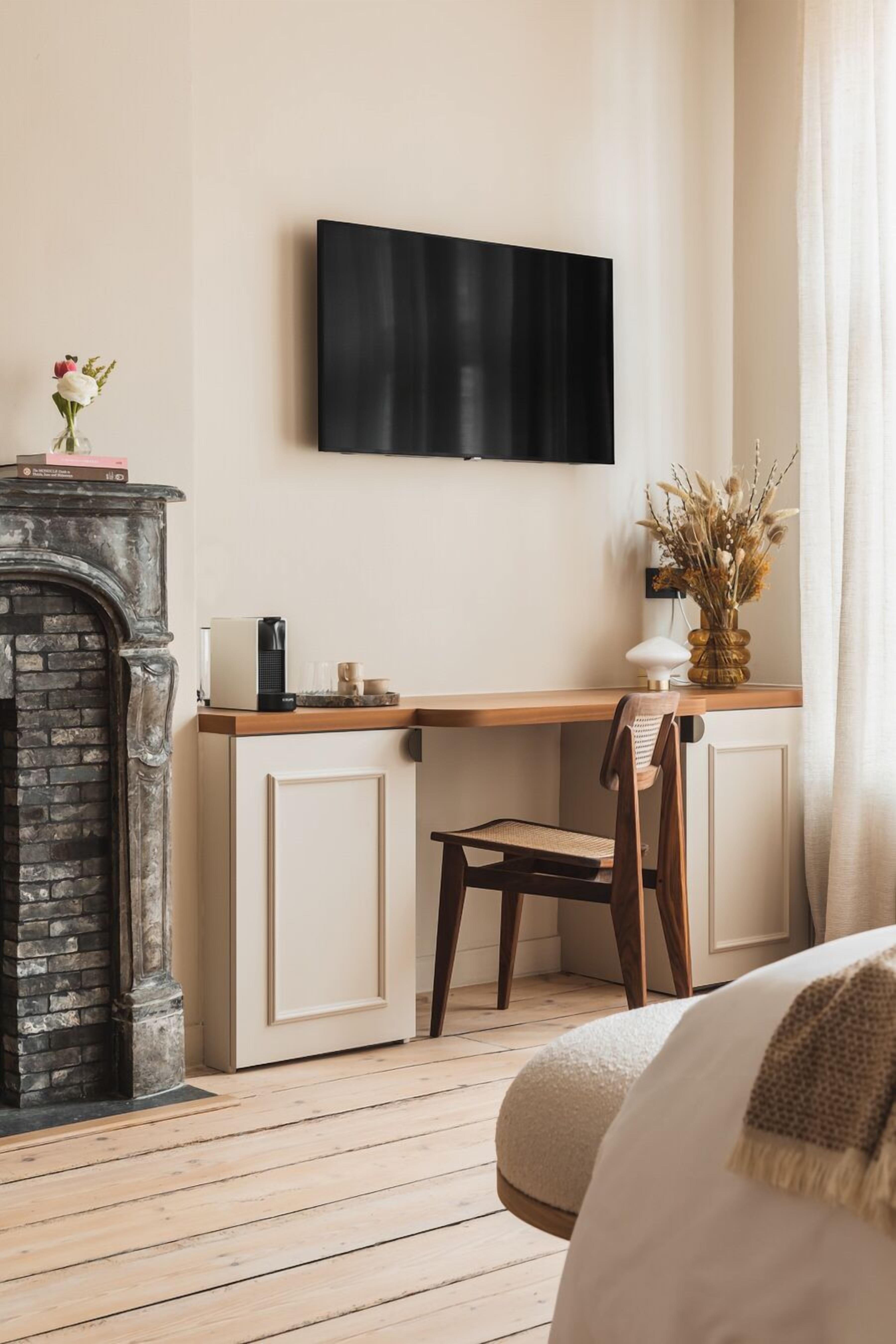A cozy room with a fireplace, a desk with a chair, a television mounted on the wall, and a window with curtains.
