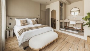Quarto com cama grande, um banquinho, piso de madeira, banheiro com espelho e uma planta.