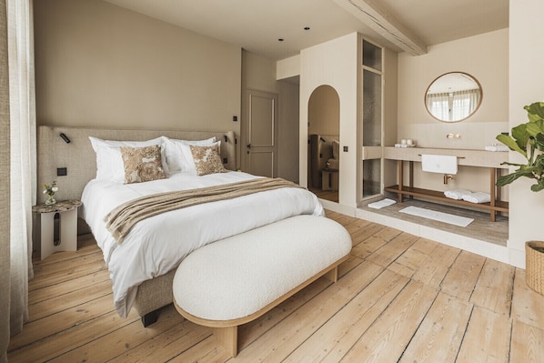A bedroom with a large bed, a bench, a wooden floor, a bathroom with a mirror, and a plant.