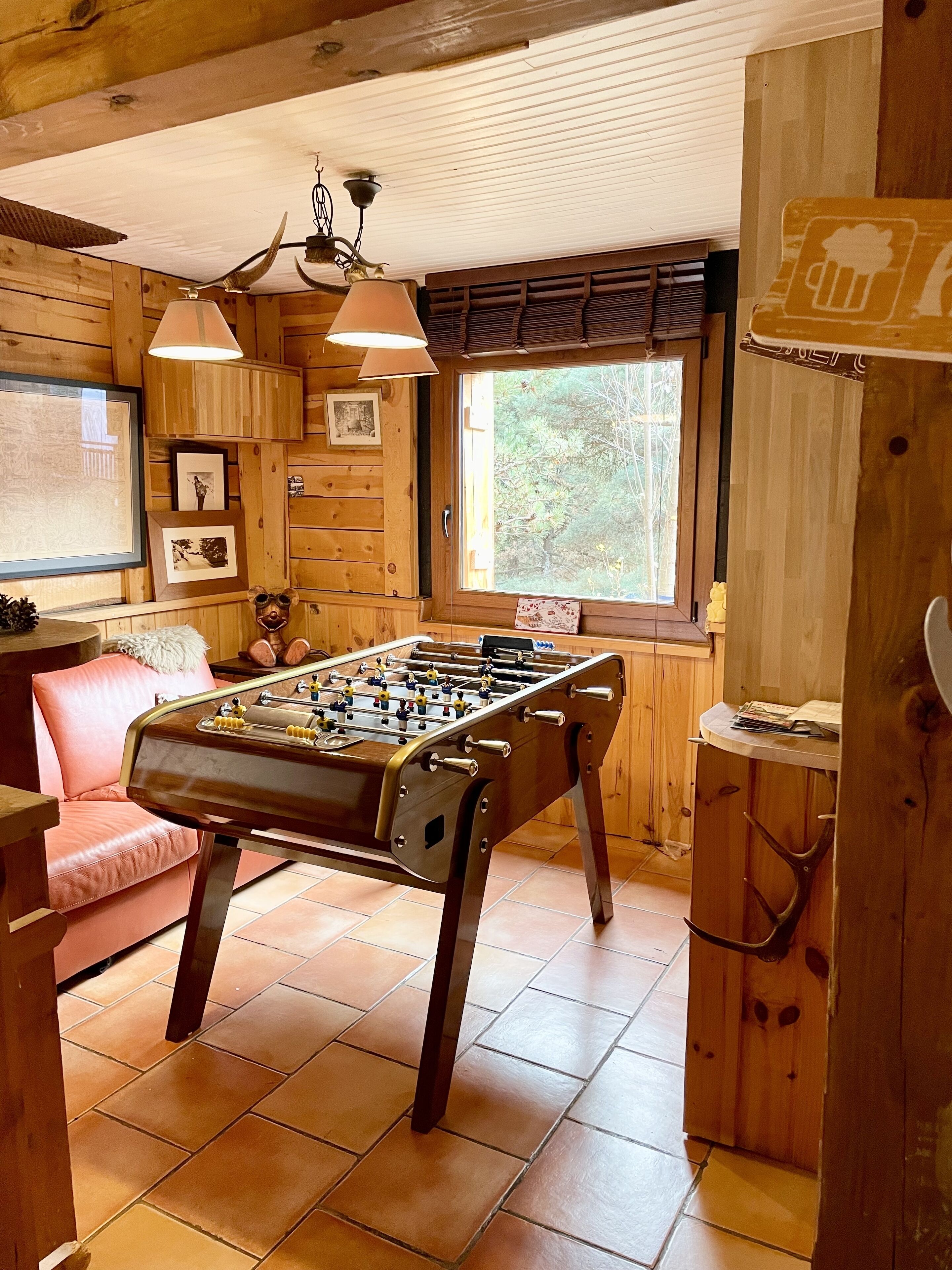Ein gemütliches Zimmer mit Kicker, Holzwänden, einem rosa Sessel und einem Fenster mit Blick auf Bäume.