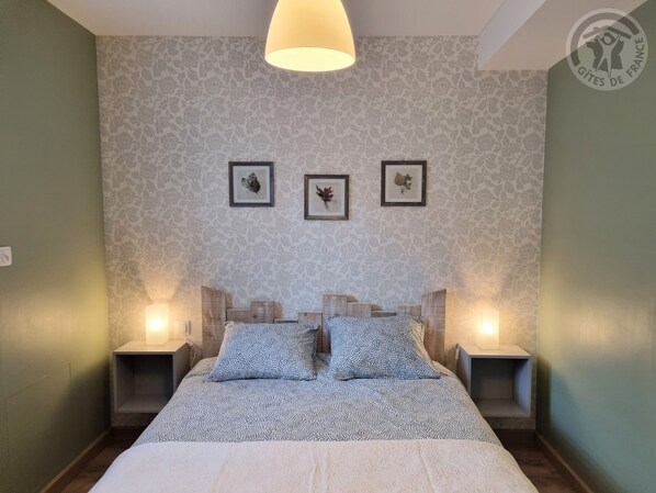 Ein Schlafzimmer mit gemustertem Tapetenbelag, einem Bett mit einer gemusterten Tagesdecke, zwei Nachttischen mit Lampen und drei gerahmten Bildern an der Wand.