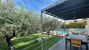 Une terrasse couverte avec une piscine, des sièges d’extérieur et une vue sur une pelouse et des arbres bien entretenus.