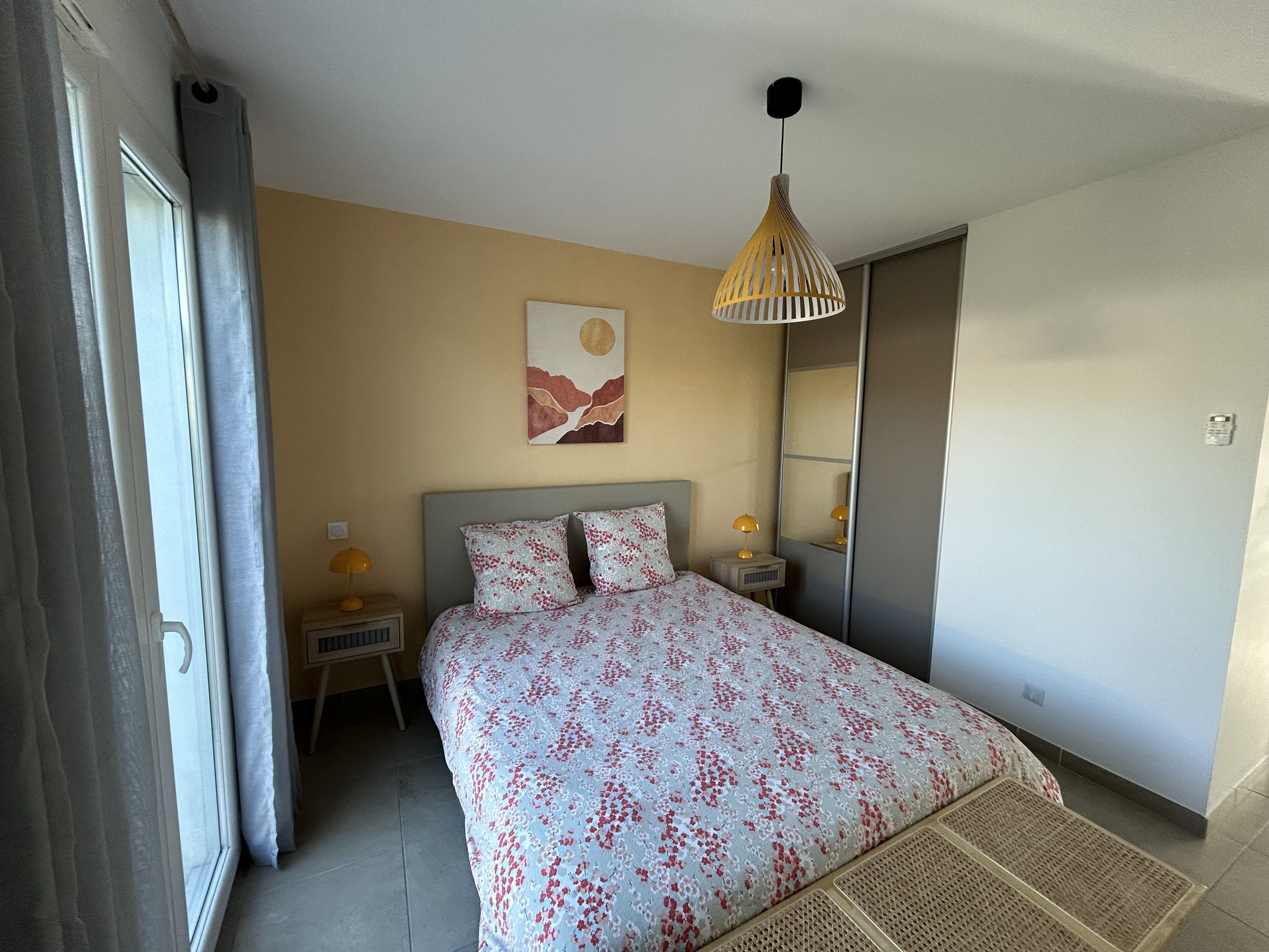 A bedroom with a bed, a bedside table with a lamp, a hanging light fixture, a wall-mounted artwork, and a door.