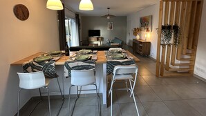 Un coin repas avec une table en bois dressée pour quatre personnes, des chaises blanches et des luminaires suspendus.