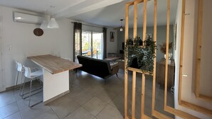 Une cuisine moderne avec un plan de travail en bois et des chaises blanches, un coin repas avec un canapé noir, et une vue sur l’extérieur grâce à de grandes fenêtres.