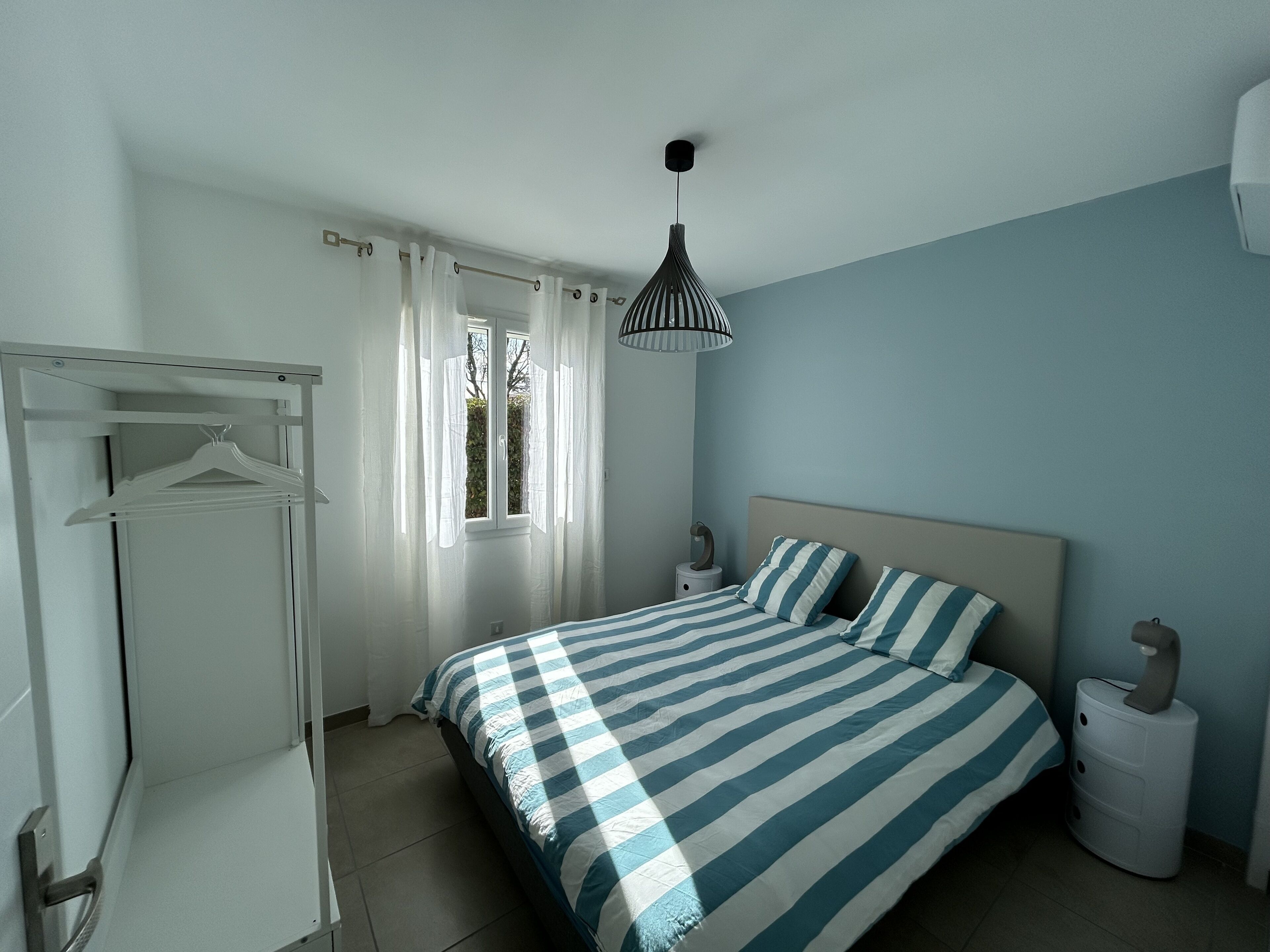 Ein Schlafzimmer mit Bett, Kleiderschrank, Nachttisch, Lampe und einem Fenster mit Vorhängen.