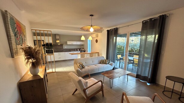 Un salon moderne avec un canapé, des fauteuils et une table basse. On aperçoit une cuisine à l’arrière-plan, avec un coin repas visible à travers une porte vitrée.