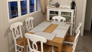 Een eetgedeelte met een houten tafel, witte stoelen, een witte kast met een klok en een raam met gordijnen.