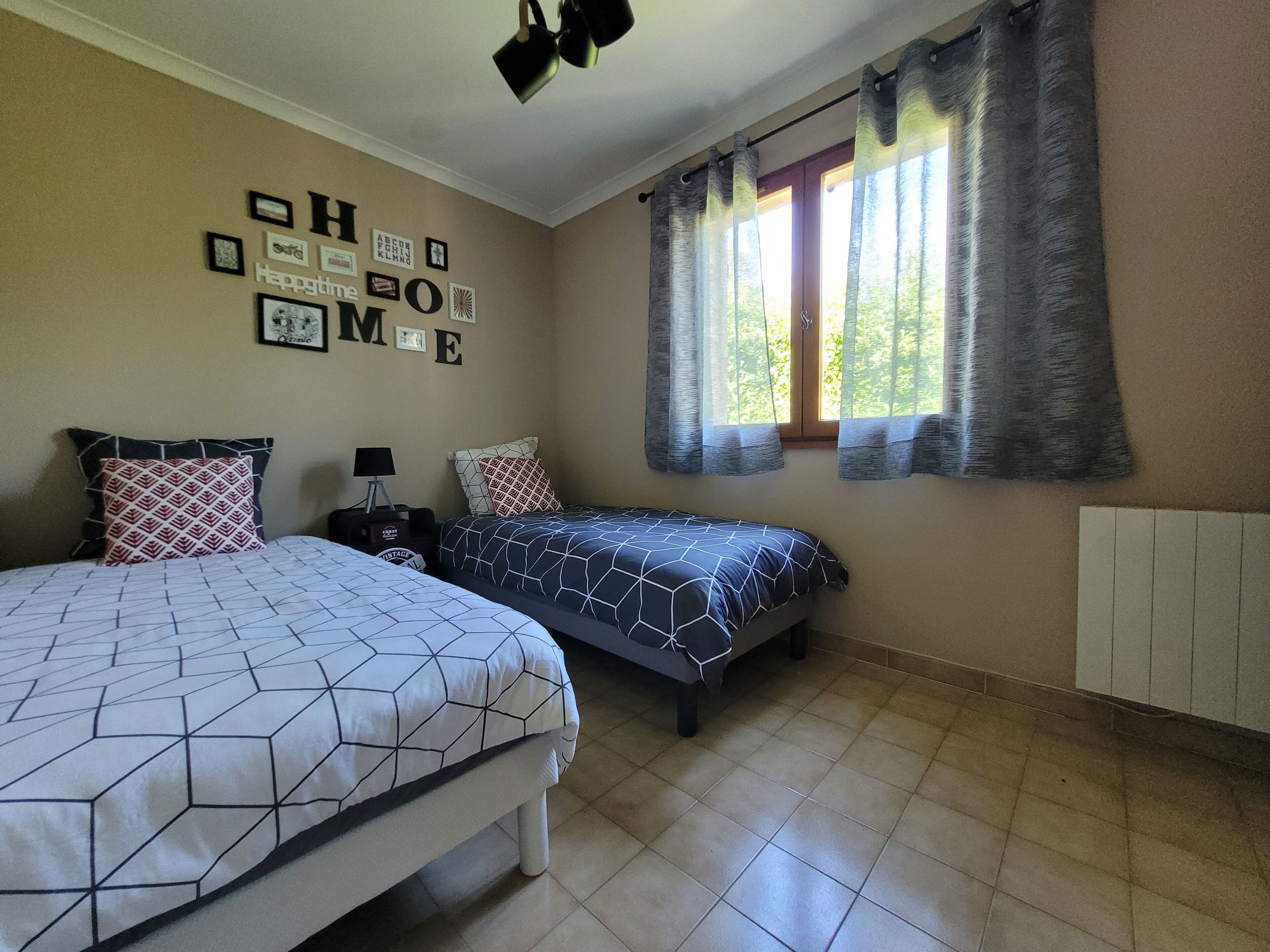 Une chambre avec deux lits, un mur orné de lettres de l’alphabet et d’une enseigne « Happytime HOME », une fenêtre avec des rideaux et un radiateur.