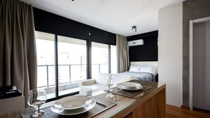 Une chambre d’hôtel moderne, dotée d’un lit, d’un coin repas avec une table dressée pour deux personnes, et de grandes fenêtres offrant une vue sur la ville.