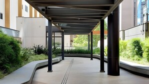 Une allée couverte, dotée d’un chemin tactile pour les personnes malvoyantes, bordée de verdure et entourée de bâtiments modernes.