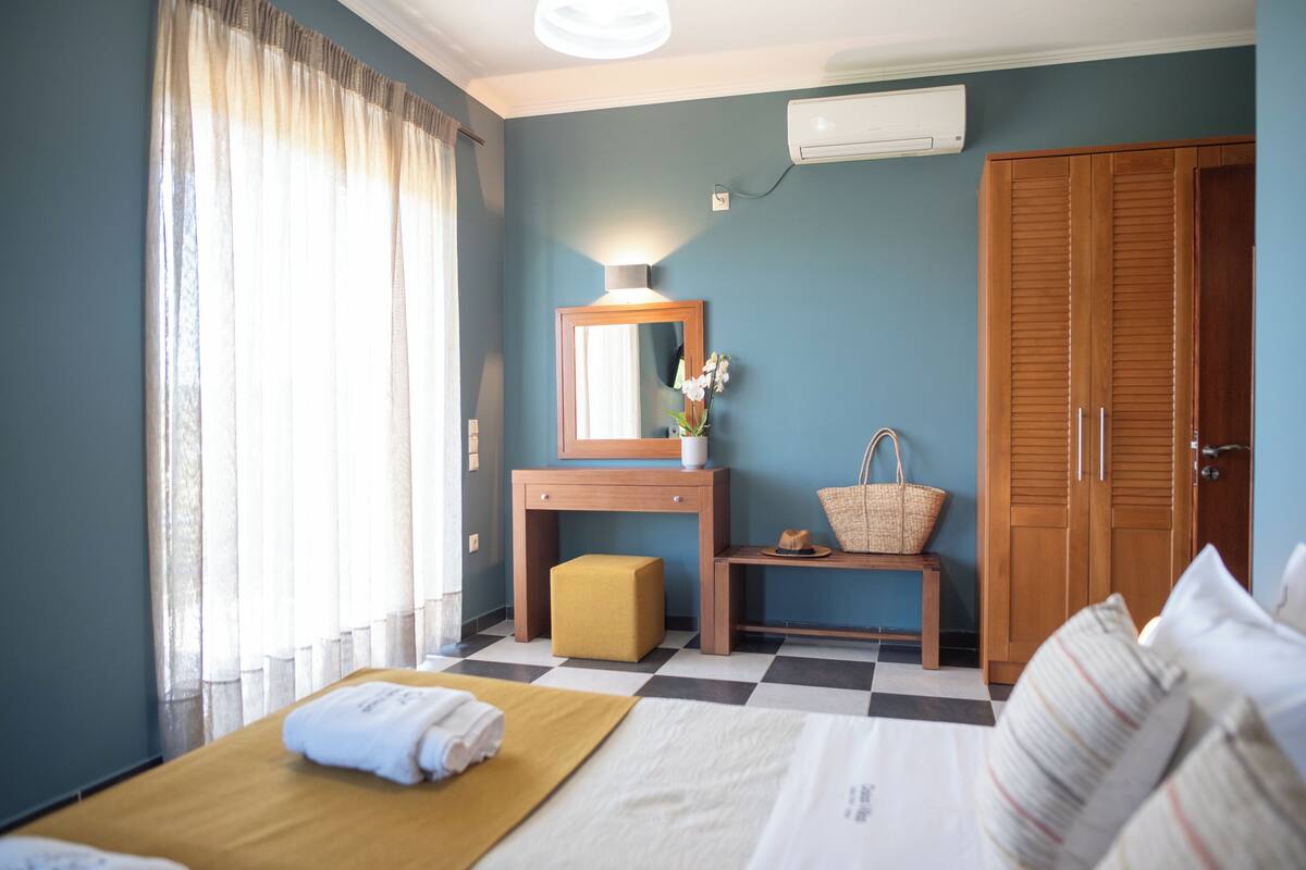 A hotel room with a bed, a desk with a mirror, a small stool, a basket, and a wardrobe.