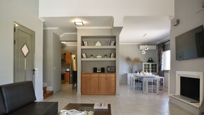 Una sala de estar moderna con sofá, una mesa de centro y un comedor con mesa y sillas. Se observa un área de cocina al fondo, con armarios y un refrigerador.