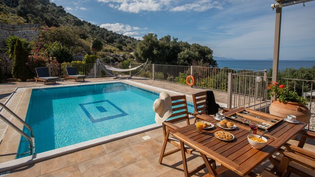 Zona de piscina con vista al mar, una hamaca y una mesa lista para el desayuno.