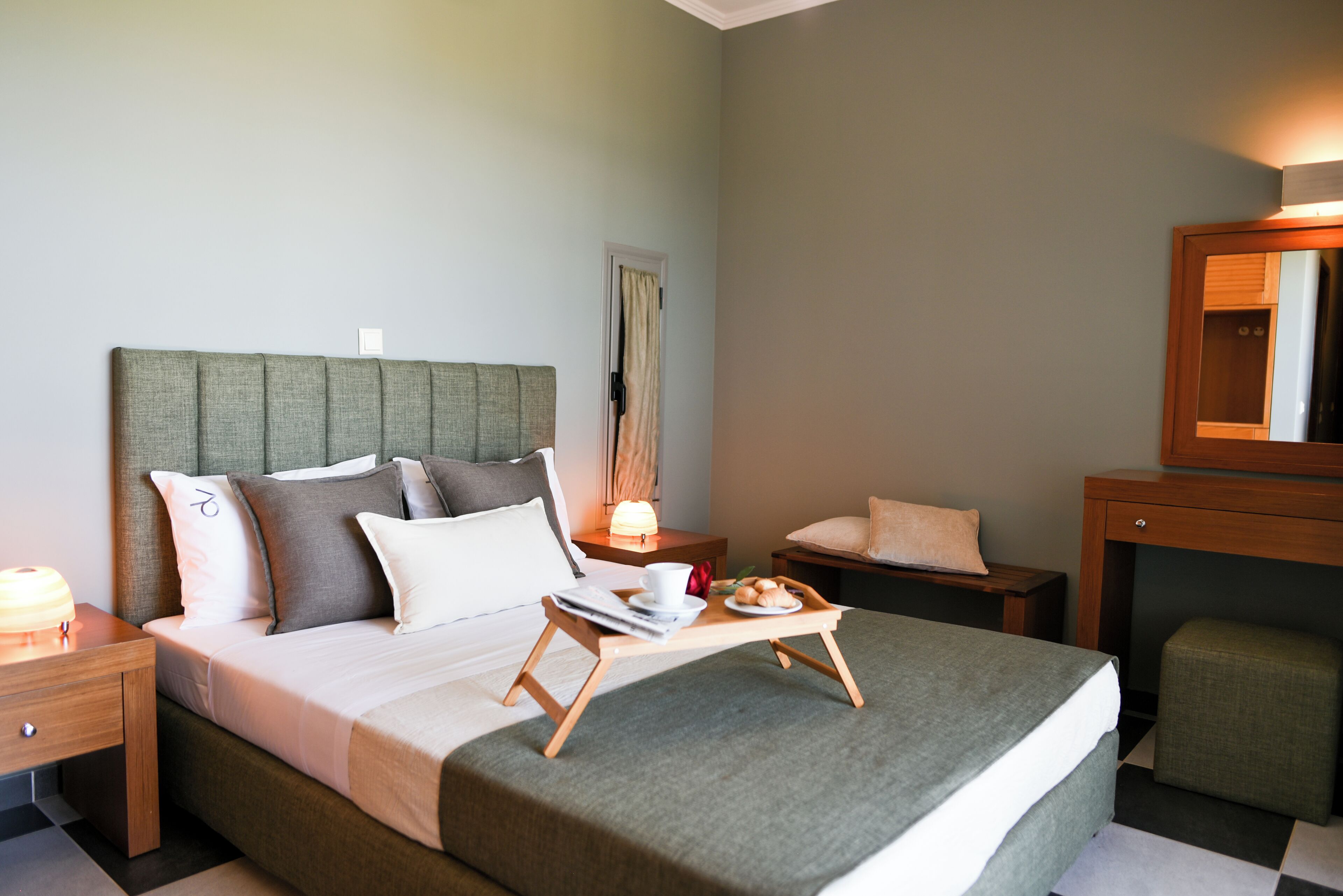 Un lit bien fait, avec des oreillers, une table de chevet sur laquelle se trouve une lampe, un plateau avec des éléments de petit-déjeuner et un miroir au-dessus d’un bureau en bois.