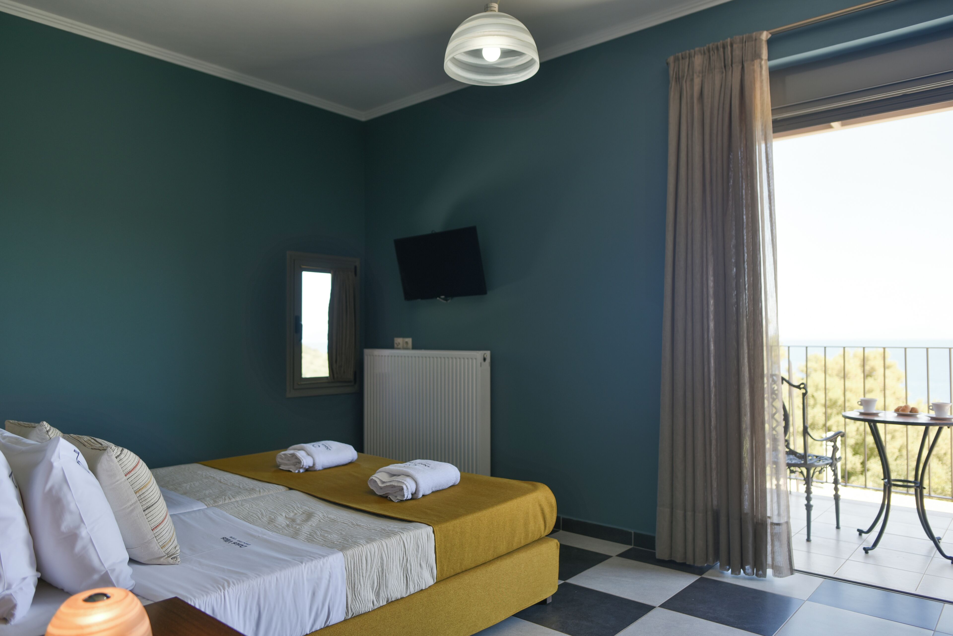 A hotel room with a bed, a television, a window with curtains, a radiator, and a balcony with a table and chair.