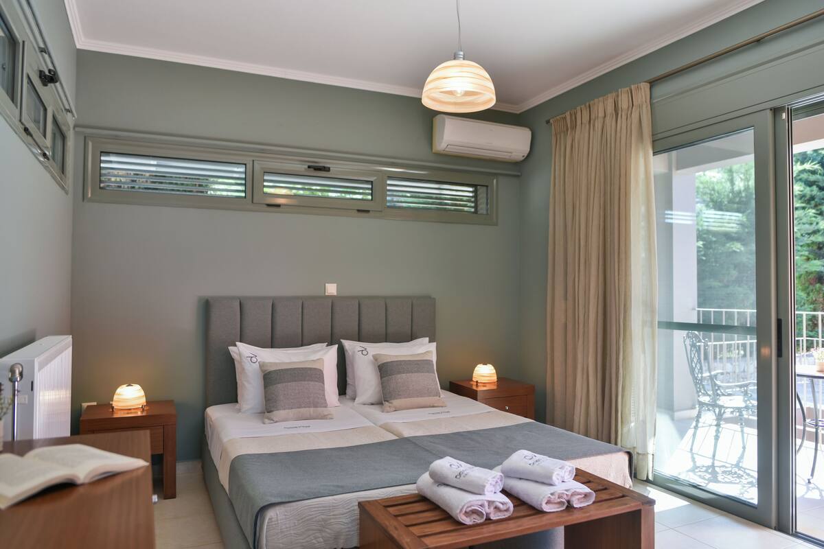 A bedroom with a large bed, two bedside tables, a bedside lamp, a window with blinds, and a view of the outdoors.