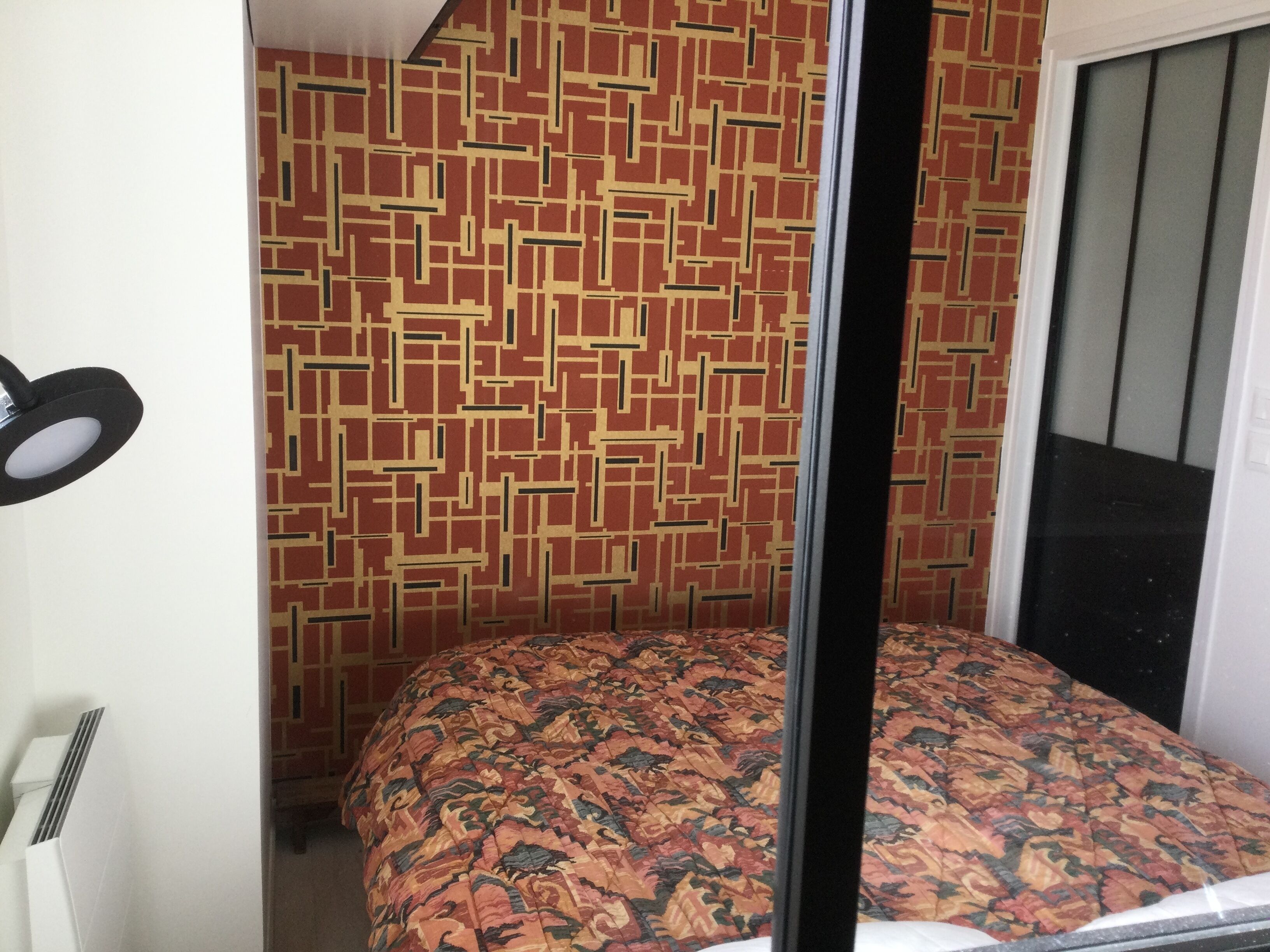 A room with a patterned wall, a bed with a floral bedspread, a black pole, and a white radiator.