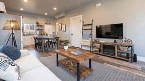 A modern living room with a sofa, coffee table, and TV. There is a kitchen area with cabinets and a dining table.