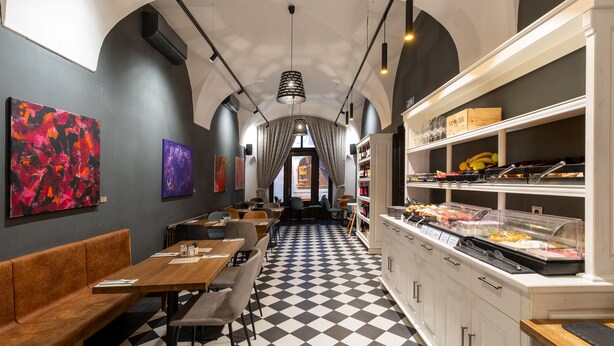 Un restaurante con piso de tablones de ajedrez, mesas y bancos de madera. Hay obras de arte en las paredes y un bufé con diferentes alimentos.