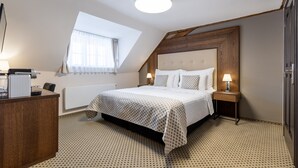 Un dormitorio con cama, mesitas de noche, escritorio, radiador, ventana con cortinas y techo inclinado.
