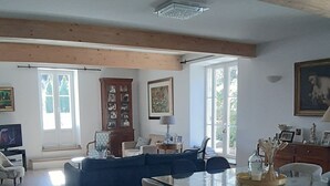 A spacious living room with a glass-top dining table, wooden beams, and a large window.