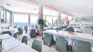 A spacious dining area with multiple tables and chairs, large windows offering a view of the sea, and a mural on the wall.