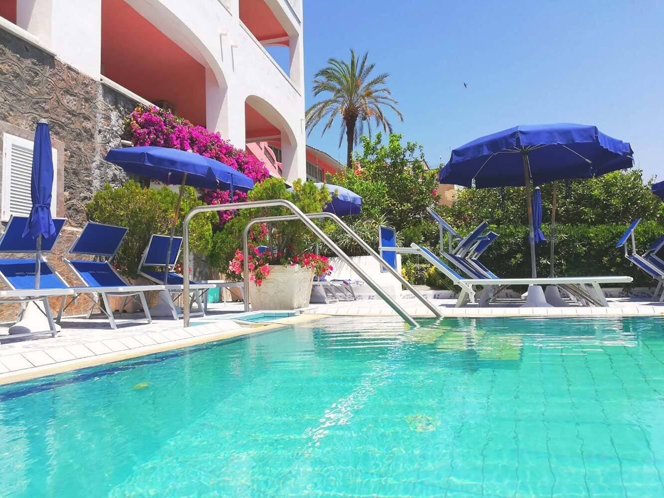A swimming pool with blue lounge chairs and umbrellas, surrounded by a building with balconies and pink flowers.