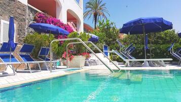 A swimming pool with blue lounge chairs and umbrellas, surrounded by a building with balconies and pink flowers.