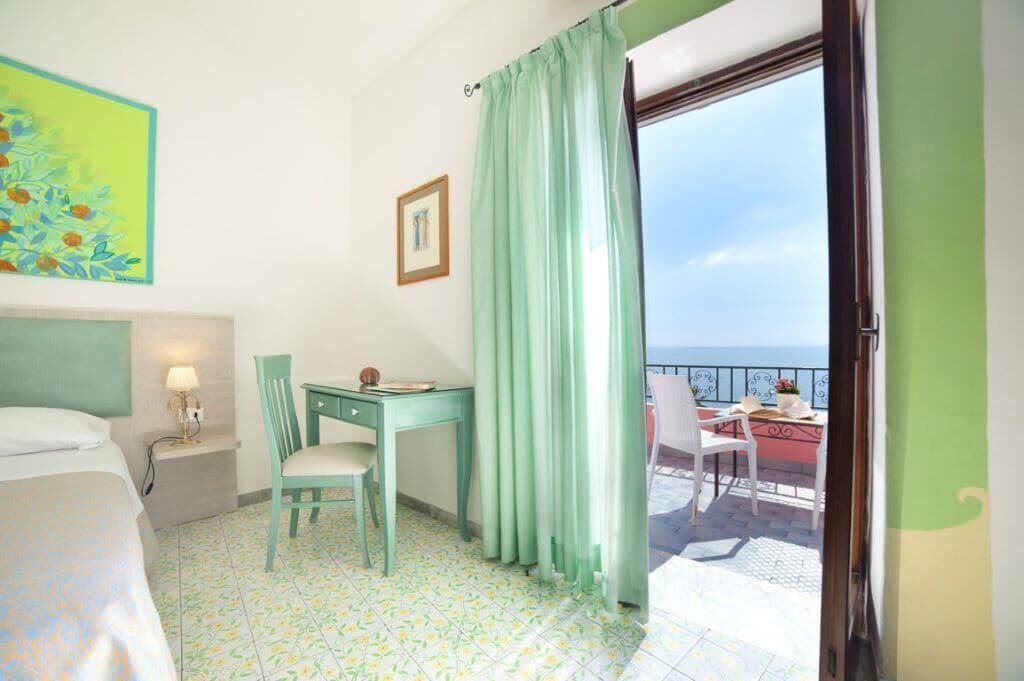 Ein Zimmer mit einem Bett, einem Schreibtisch und einem Sessel, ein Fenster mit Meerblick und ein Balkon mit Tisch und Stühlen.