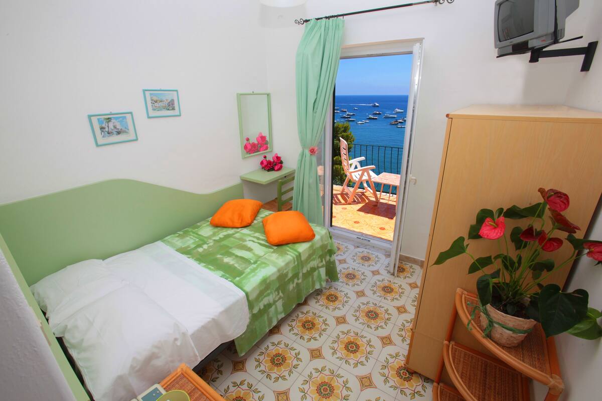Ein Schlafzimmer mit einem Bett, einem kleinen Tisch mit Spiegel, einer Topfpflanze und Blick auf Boote auf dem Wasser durch eine offene Tür.