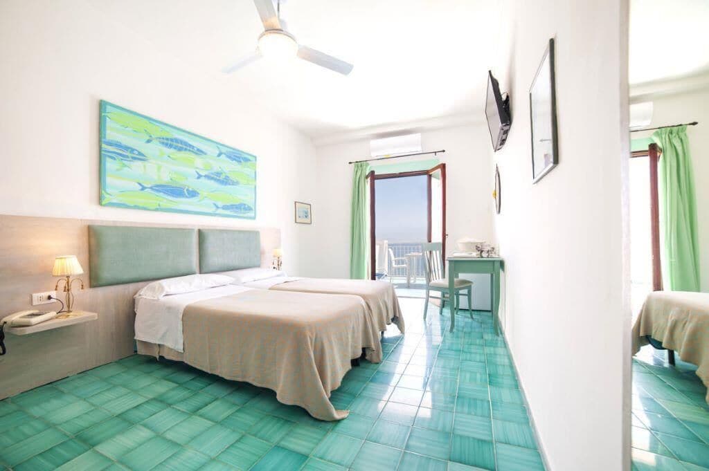 A hotel room with two beds, a desk, a chair, a balcony, and a large window.