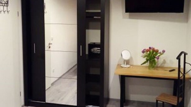 A room with a black wardrobe, a wooden desk, a chair, a TV mounted on the wall, and a vase with flowers on the desk. - Modern vacation apartment in a quiet location (Meißner)